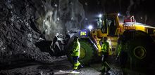 Foto: tre tunnelarbeidere i arbeidsklær og hjelm går mot en mørk fjellvegg, en gravemaskin og en tunnelvegg sprayet med betong i bakgrunnen.